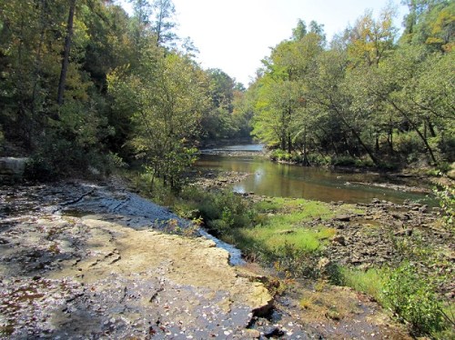 A serene river scene surrounded by trees, with rocky banks and calm water reflecting the clear sky.