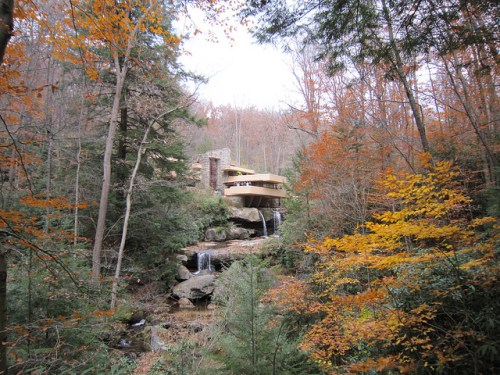 A modern house perched on a rocky outcrop, surrounded by autumn foliage and cascading waterfalls in a forested area.