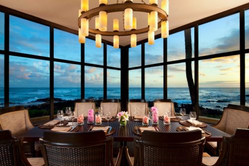 Elegant dining room with a large table, surrounded by windows showcasing a sunset over the ocean.