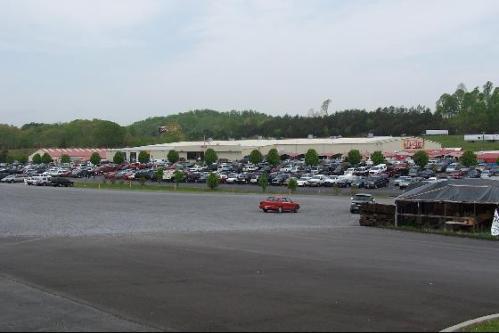 A large parking lot filled with cars in front of a retail building, surrounded by greenery and a cloudy sky.