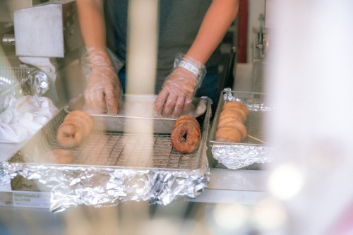 A person in gloves prepares donuts on a tray, with freshly fried donuts visible in the foreground.
