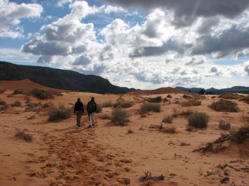 Two people walk through a sandy desert landscape under a cloudy sky, with distant mountains in the background.
