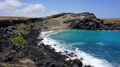 A scenic view of a rocky coastline with turquoise waters and a sandy beach under a blue sky with scattered clouds.