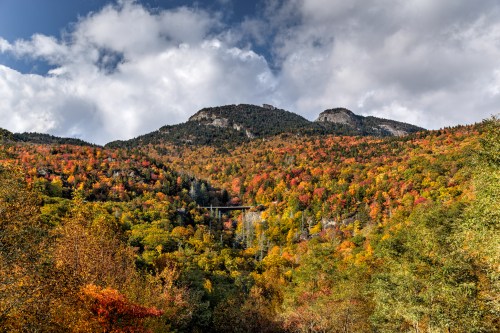 A vibrant autumn landscape with colorful trees, mountains, and a cloudy sky.
