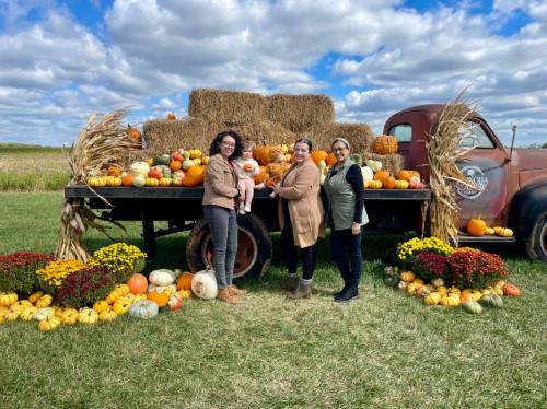 Three women and a baby pose in front of a vintage truck filled with pumpkins and hay bales on a sunny day.