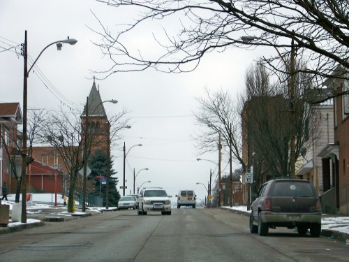 A snowy street scene with bare trees, parked cars, and a clock tower in the background under a gray sky.