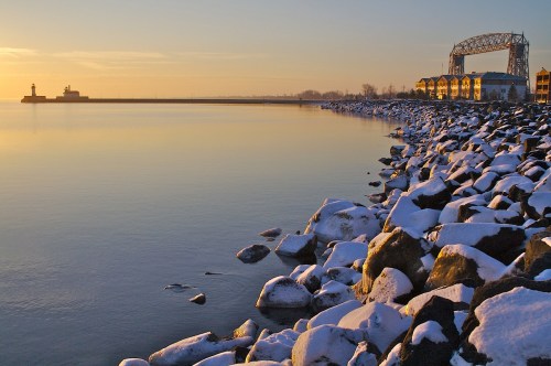 A serene lakeside view at sunrise, featuring snow-covered rocks and a distant lighthouse against a calm water surface.