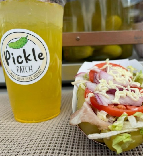 A refreshing drink next to a loaded sandwich with lettuce, tomatoes, onions, and mayonnaise, served in a pickle-shaped container.
