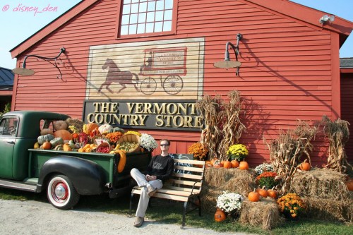 A person sits on a bench beside a vintage truck filled with pumpkins and autumn decorations in front of a red barn.