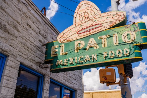A vintage sign for "El Patio Mexican Food" featuring a sombrero, against a blue sky with fluffy clouds.