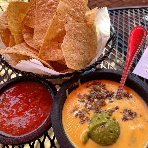 A bowl of cheesy dip with ground meat and guacamole, served with a side of tortilla chips and salsa on a black table.