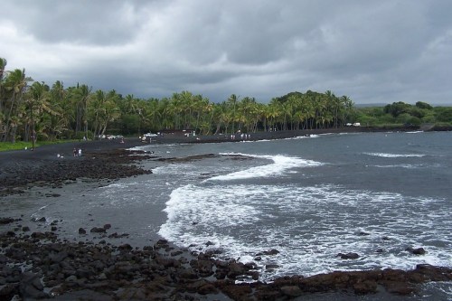 A scenic view of a black sand beach with palm trees and cloudy skies, waves gently lapping at the shore.