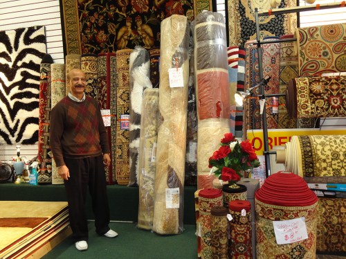 A man stands beside rolled rugs in a shop, surrounded by various patterned carpets and decorative items.