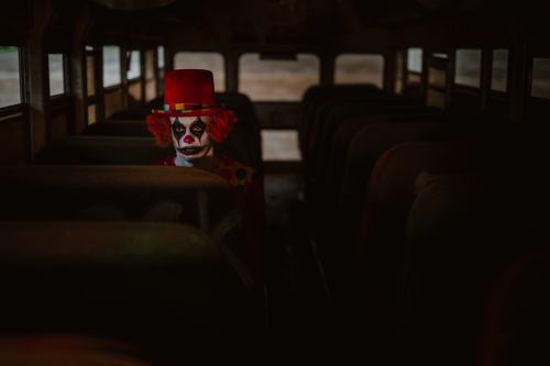 A clown in a red hat sits alone on a dark, empty bus, surrounded by worn seats.