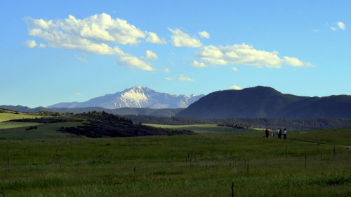 A scenic landscape featuring green fields, distant mountains, and a clear blue sky with a few clouds. People walk in the distance.