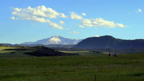 A scenic landscape featuring green fields, distant mountains, and a clear blue sky with a few clouds. People walk in the distance.