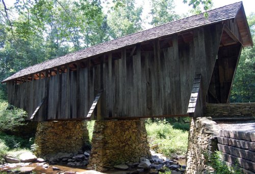 A rustic wooden covered bridge spans a small stream, surrounded by lush greenery and trees.