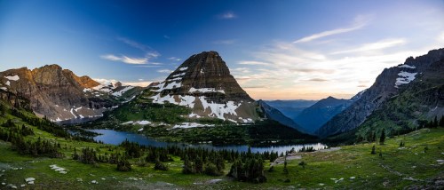 A panoramic view of a mountain landscape with snow-capped peaks, a serene lake, and lush green valleys at sunset.