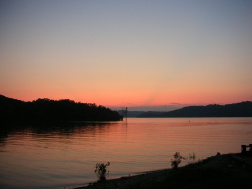A serene lake at sunset, with soft pink and orange hues reflecting on the water and silhouettes of distant hills.