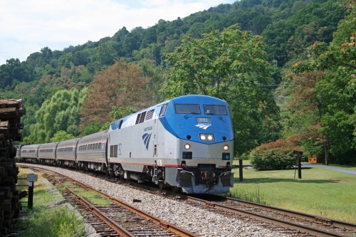 An Amtrak train travels along a scenic route, surrounded by lush green trees and hills.