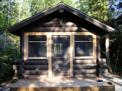 A rustic log cabin with a wooden porch, surrounded by trees and dappled sunlight.