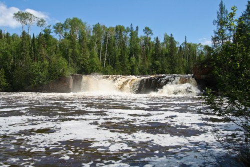A scenic waterfall cascading over rocks, surrounded by lush green trees and a blue sky.