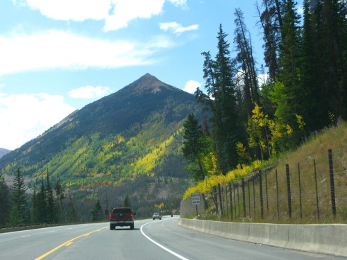 A scenic mountain road with a view of a peak and colorful trees under a blue sky with clouds.