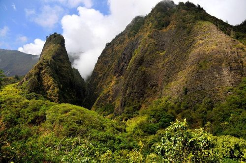 Lush green mountains rise steeply under a blue sky with scattered clouds, creating a dramatic natural landscape.