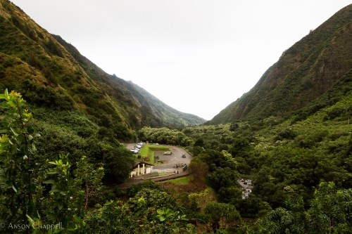 A lush green valley surrounded by mountains, with a parking area and a winding road visible below.