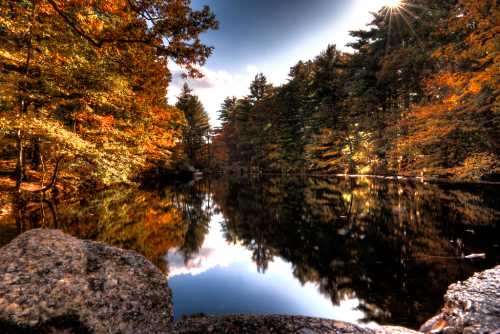 A serene lake surrounded by vibrant autumn trees, reflecting the colorful foliage and a bright sunburst in the sky.