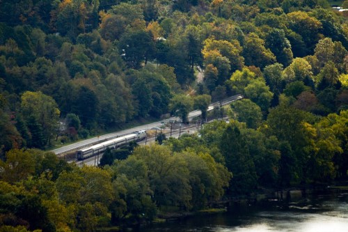 A train travels through a colorful forested area beside a river, showcasing vibrant autumn foliage.