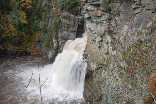 A waterfall cascading over rocky cliffs into a river, surrounded by autumn foliage and rugged terrain.