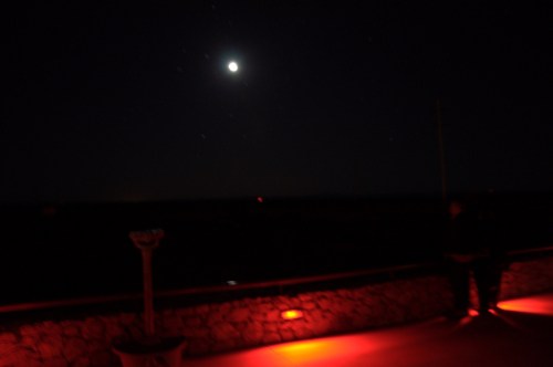 A dimly lit outdoor scene at night with a bright moon and a person sitting near a stone wall illuminated by red light.