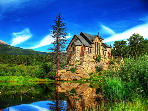 A stone church surrounded by greenery and a calm pond, reflecting the blue sky and clouds in the background.