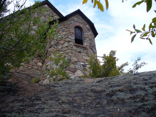 A stone building with a window, surrounded by greenery and rocky terrain under a cloudy sky.