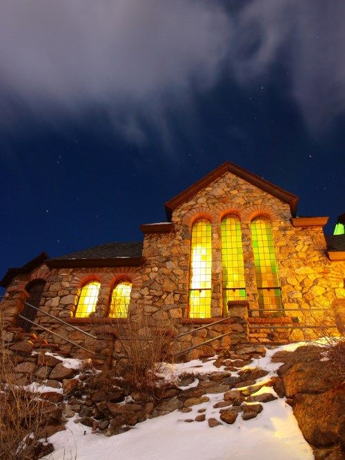 A stone building illuminated at night, with glowing windows and a starry sky above, surrounded by snow.