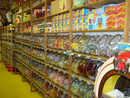 A colorful candy store with glass jars filled with various candies on wooden shelves against a bright yellow floor.