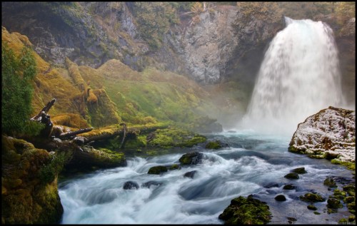 A serene waterfall cascades into a misty river, surrounded by lush greenery and moss-covered rocks.