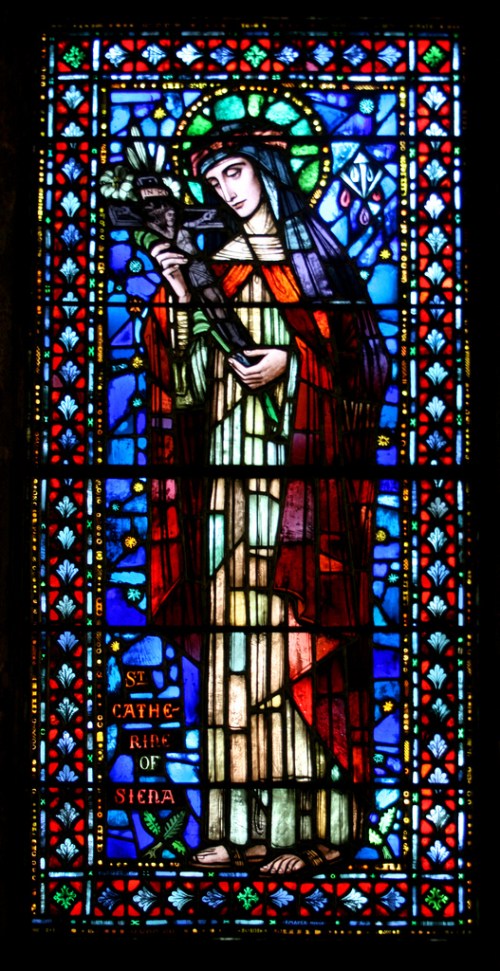 Stained glass window depicting St. Catherine of Siena, holding a cross, surrounded by vibrant blue and red colors.