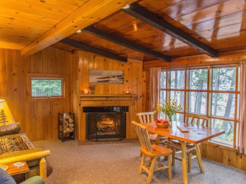 Cozy wooden cabin interior with a fireplace, dining table, and large windows overlooking a forested area.