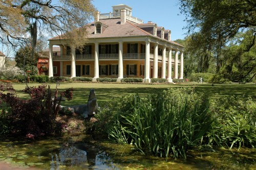A grand, two-story mansion with columns, surrounded by lush greenery and a pond in the foreground.