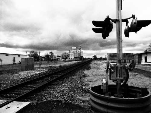 Black and white image of a railway crossing signal beside tracks, with a cloudy sky and industrial buildings in the background.