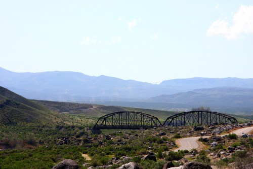 A metal bridge spans a valley, surrounded by rolling hills and mountains under a clear blue sky.