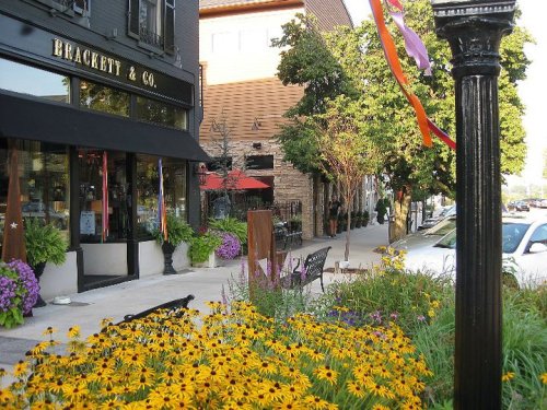 A vibrant street scene featuring a flower-filled sidewalk, a shop front, and colorful ribbons hanging from a lamppost.