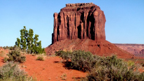 A towering red rock formation rises above a desert landscape, surrounded by sparse vegetation and clear blue skies.