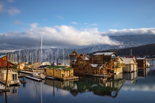 Colorful houseboats and sailboats reflect on calm water under a blue sky with clouds and mountains in the background.