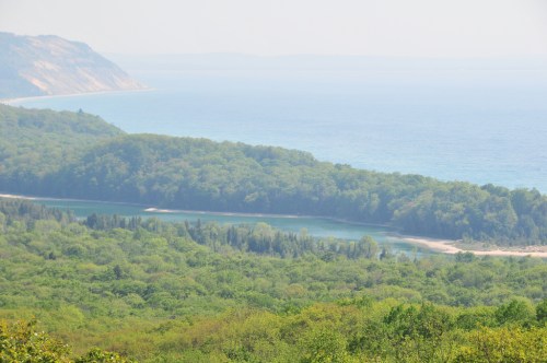 A scenic view of lush green forests meeting a calm blue lake and distant hills under a hazy sky.