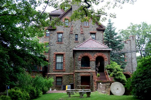 A large stone building with a red-tiled roof, surrounded by greenery and featuring a stone bench in the foreground.