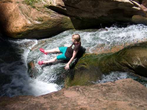 A person in a brown shirt and teal shorts sits on a rock in a flowing stream, enjoying the water.