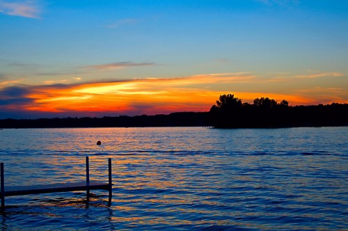 A serene sunset over a calm lake, with vibrant orange and blue hues reflecting on the water. A small dock is visible.